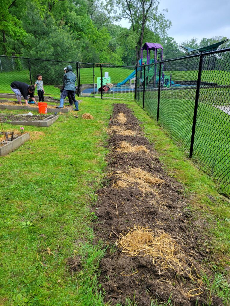 Photo shows HEARTH's community garden. Families are seen digging in planters in the background. A row of dirt is freshly piled on the right side of the garden.